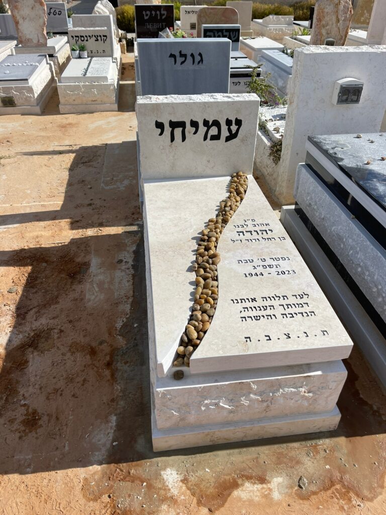 מצבה לבנה מעוצבת בבית העלמין עם שביל אבנים וכתובת בעברית.