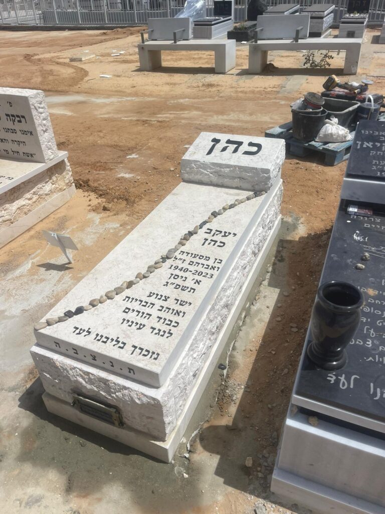 מצבת אבן לבנה מעוצבת עם כיתוב בעברית ושורת אבנים בבית עלמין