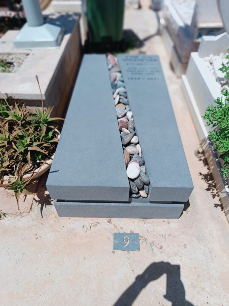 מצבה מודרנית אפורה עם חריץ מלא אבנים בבית עלמין