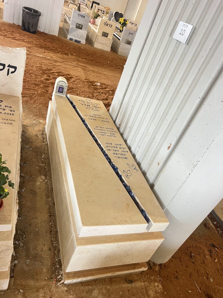 מצבות מאבן מסודרות במחסן תצוגה עם רצפת אדמה ולוחות זיהוי.